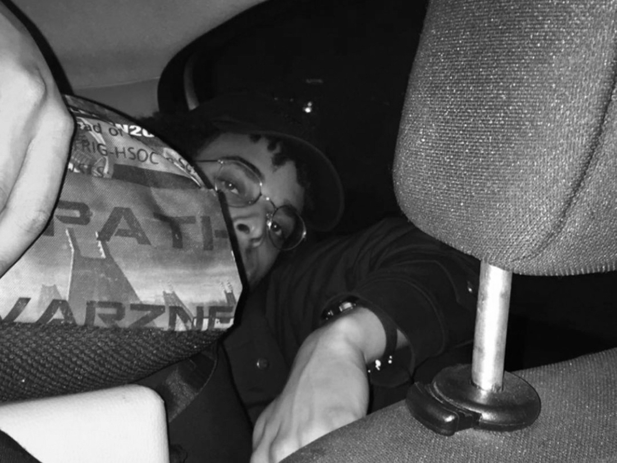 Schwarz-Weiss Aufnahme von Tapiwa Svosve, welcher von der Rückback eines Autos zwischen den Kopfstützen in die Kamera linst.